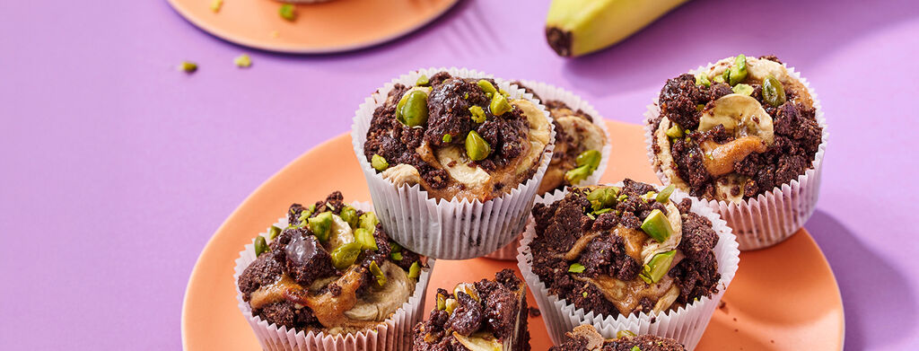 Chocolate banana cupcakes arranged on a round plate. The cupcakes are topped with dark chocolate crumble, banana slices, and chopped green pistachios. A bunch of ripe yellow bananas with a visible Dole sticker is placed in the upper right corner.