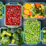 Mehrere Behälter mit gefrorenen Lebensmitteln auf einem Tisch. In den Behältern ist verschiedene Obst und Gemüse zu sehen. Wie zum Beispiel Erbsen, Rosenkohl und Himbeeren.