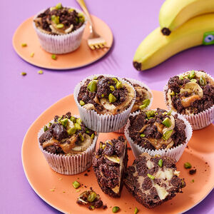 Chocolate banana cupcakes arranged on a round plate. The cupcakes are topped with dark chocolate crumble, banana slices, and chopped green pistachios. A bunch of ripe yellow bananas with a visible Dole sticker is placed in the upper right corner.