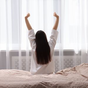 Eine Frau sitzt auf einem Bett und dehnt sich. Ihre Arme sind nach oben gestreckt.