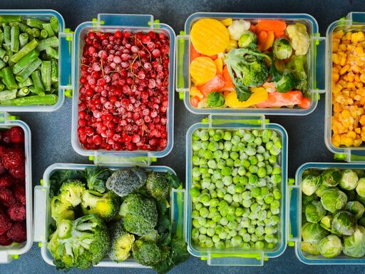 Mehrere Behälter mit gefrorenen Lebensmitteln auf einem Tisch. In den Behältern ist verschiedene Obst und Gemüse zu sehen. Wie zum Beispiel Erbsen, Rosenkohl und Himbeeren.
