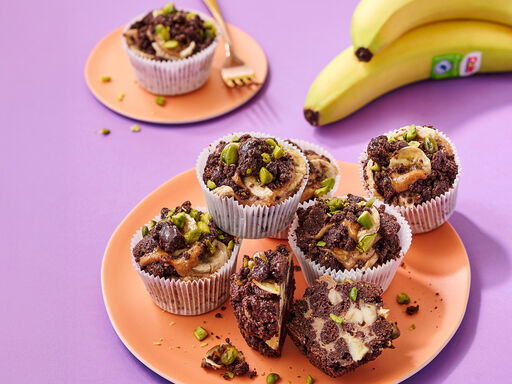 Chocolate banana cupcakes arranged on a round plate. The cupcakes are topped with dark chocolate crumble, banana slices, and chopped green pistachios. A bunch of ripe yellow bananas with a visible Dole sticker is placed in the upper right corner.