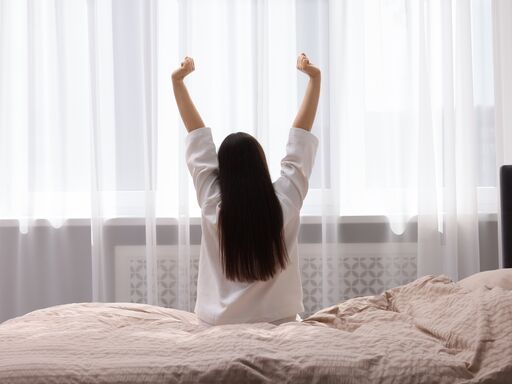 Eine Frau sitzt auf einem Bett und dehnt sich. Ihre Arme sind nach oben gestreckt.