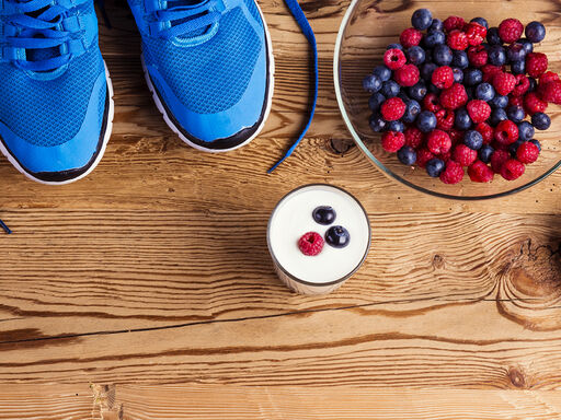 Image shows blue running shoes on a wooden floor. Next to the shoes there are 3 bananas, one bowl with mixed berries, an Avocado cut in half, a yoghurt with berries, 2 muesli bars and 1,5 oranges.